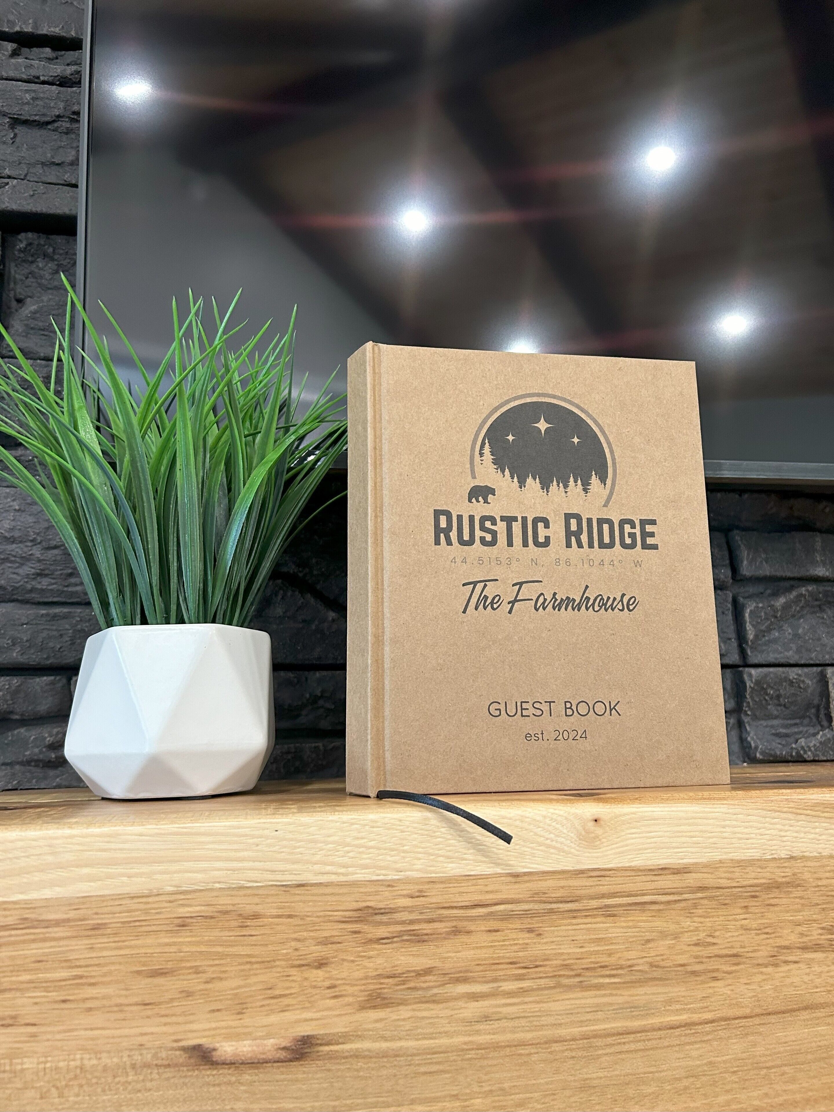 A guest book titled 'Rustic Ridge The Farmhouse' with a mountain and stars logo, placed on a wooden surface next to a potted plant.
