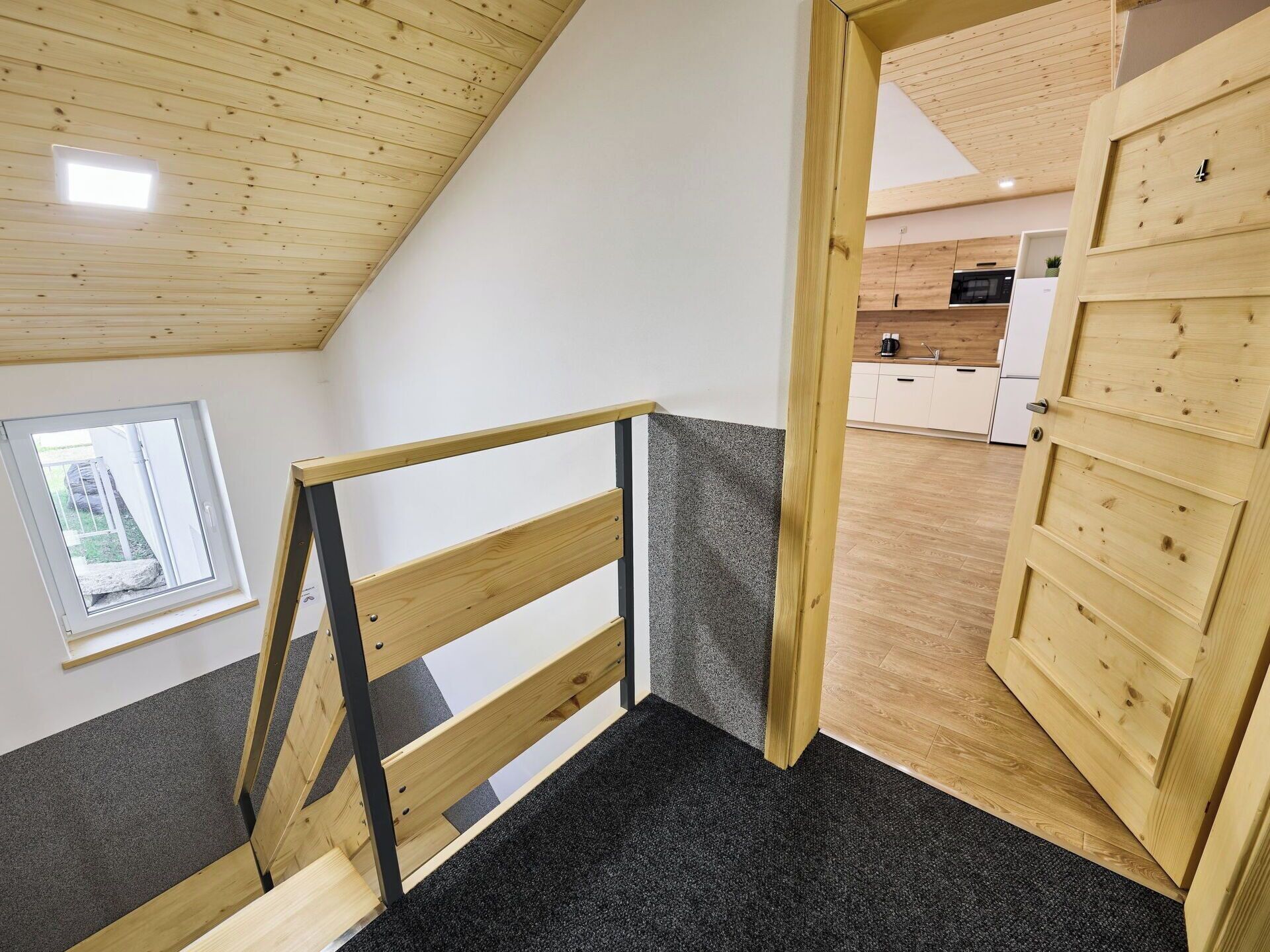 Una escalera de madera con barandilla de metal que conduce a una habitación con cocina y ventana.