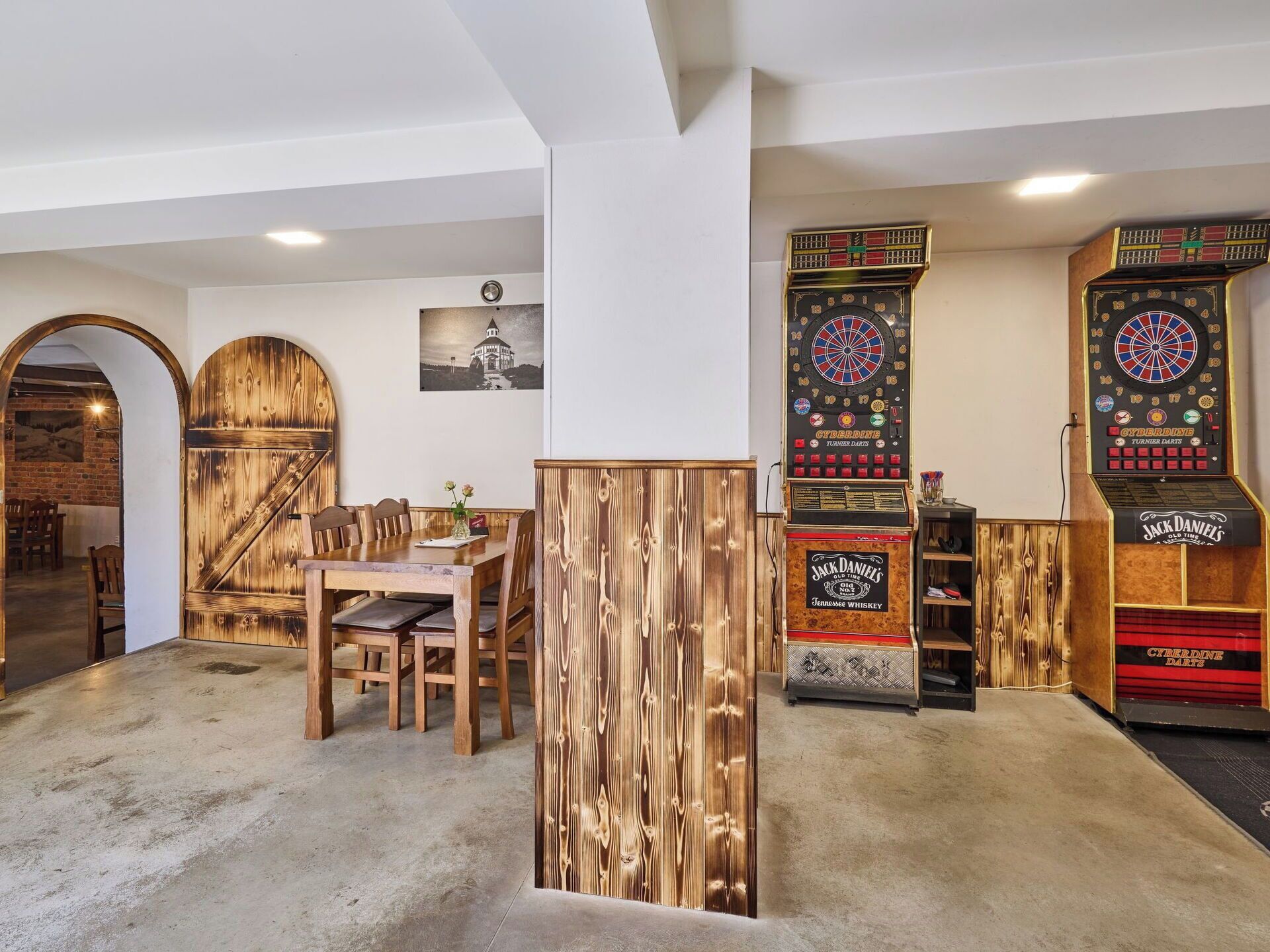 A room with a wooden dining table, chairs, a dartboard, and a jukebox.