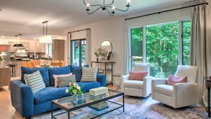 Ein Wohnzimmer mit einem blauen Sofa, einem weißen Sessel und einem Glastisch. Über dem Sofa hängt eine Chandelier und ein großes Fenster bietet Blick auf Bäume.