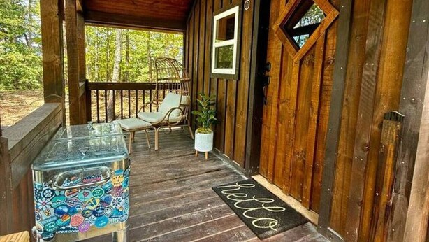 Eine hölzerne Veranda mit einem Metalltisch, zwei Stühlen, einem Blumentopf und einer Fußmatte.