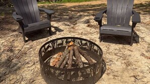 Two Adirondack chairs facing a fire pit in a natural setting with trees in the background. - Adventurer’s Nook | Tiny Home w/ Hot Tub & Firepit (Campton)