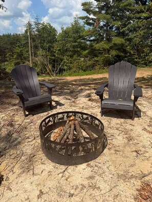 Two Adirondack chairs facing a fire pit in a natural setting with trees in the background. - Adventurer’s Nook | Tiny Home w/ Hot Tub & Firepit (Campton)