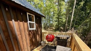 A wooden deck with a round table and two woven chairs, a barbecue grill, and a small shed. - Adventurer’s Nook | Tiny Home w/ Hot Tub & Firepit (Campton)