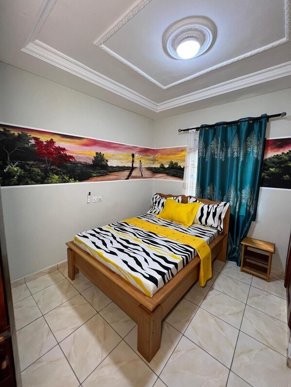 A bedroom with a bed, a small bedside table, a wall painting, and a ceiling light. - Appartment and studios in kribi. (Kribi)