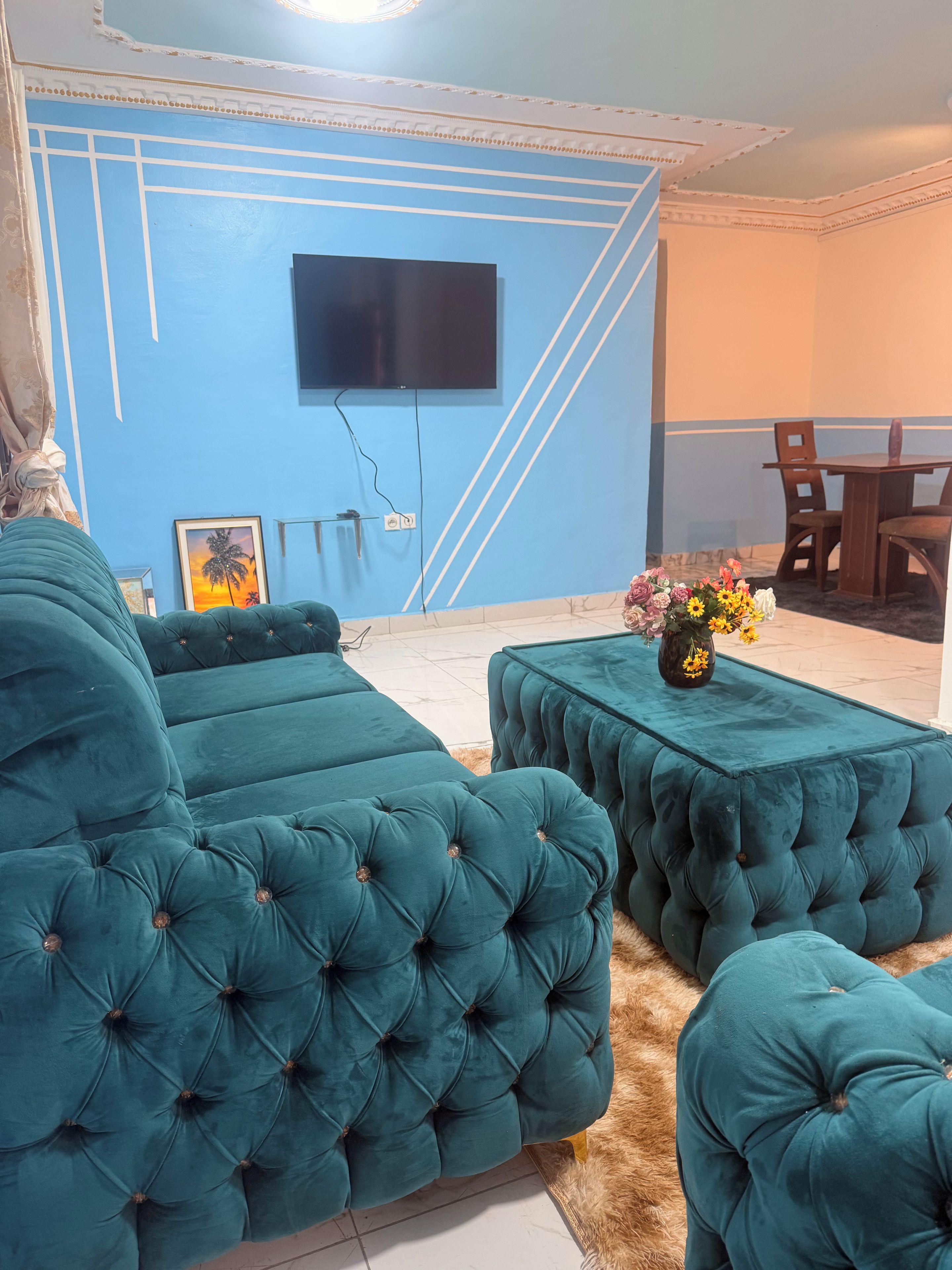 A living room with teal upholstered sofas and ottomans, a flat-screen TV mounted on the wall, and a dining area with wooden furniture.