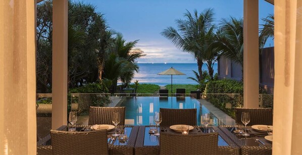 Una piscina infinita con vista al océano, rodeada de palmeras y un área de comedor con mobiliario de mimbre y mesas de vidrio.