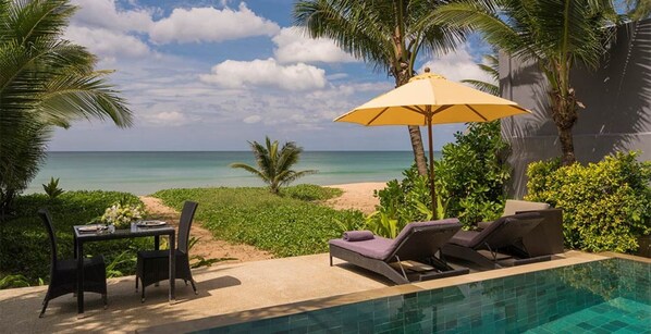 Zona junto a la piscina con vista a la playa y al océano, una mesa con sillas y sillones de descanso con sombrilla.