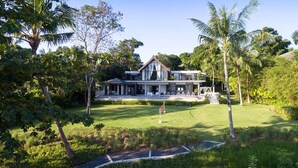 Une maison moderne à deux étages, dotée d’une grande pelouse à l’avant et entourée d’une végétation tropicale.