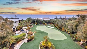 Campo de golf con vista al océano al atardecer, rodeado de vegetación exuberante y algunos edificios.