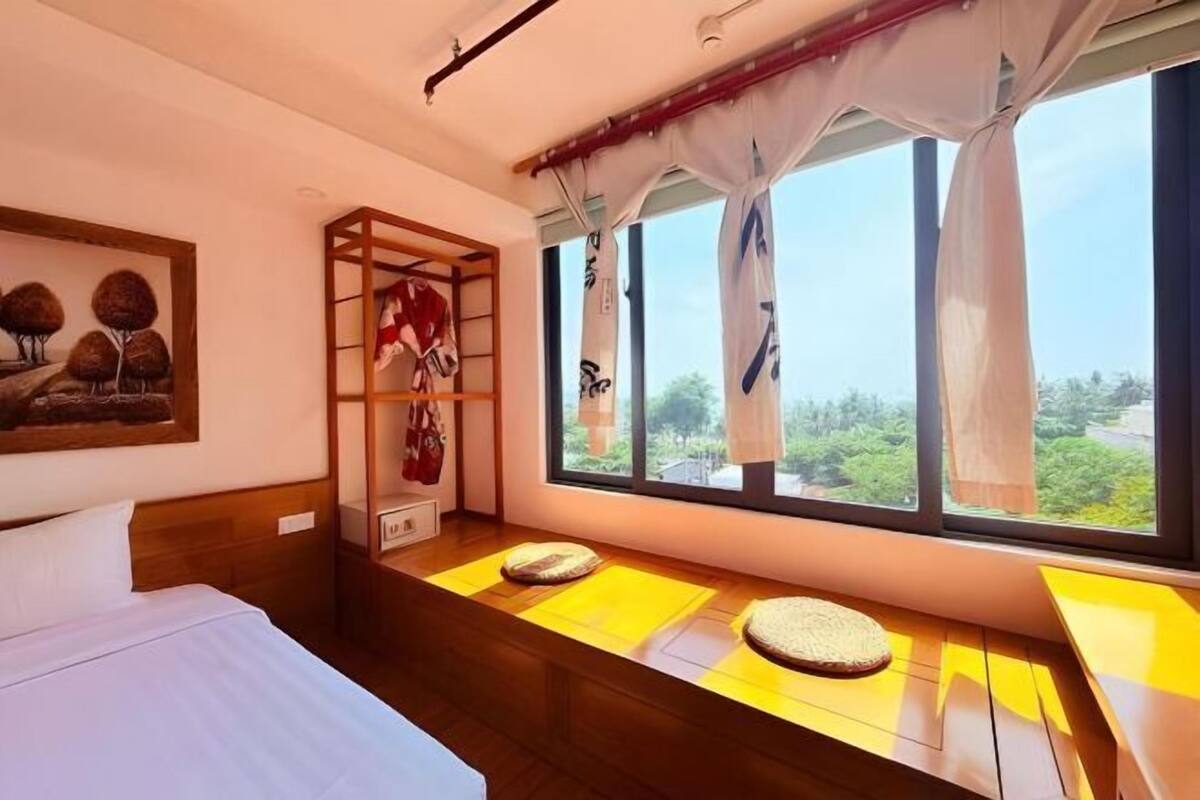 A hotel room with a bed, a wooden bench with cushions, a shelf with a red garment, and a window with curtains.