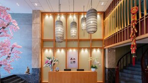 Un mostrador de recepción con detalles de madera, múltiples lámparas colgantes, una escalera y un árbol decorativo con flores rosadas.