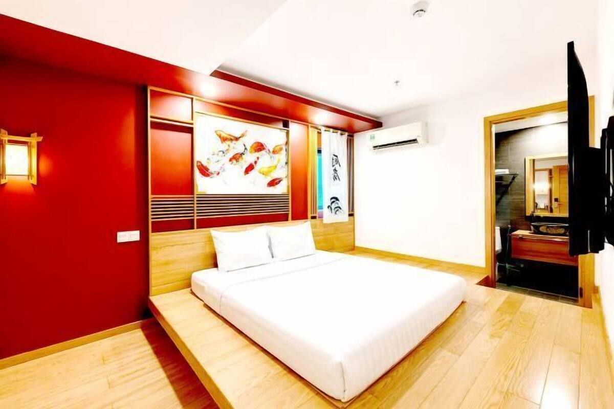 A modern hotel room with a large bed, wooden flooring, a red accent wall, and a ceiling-mounted air conditioning unit.