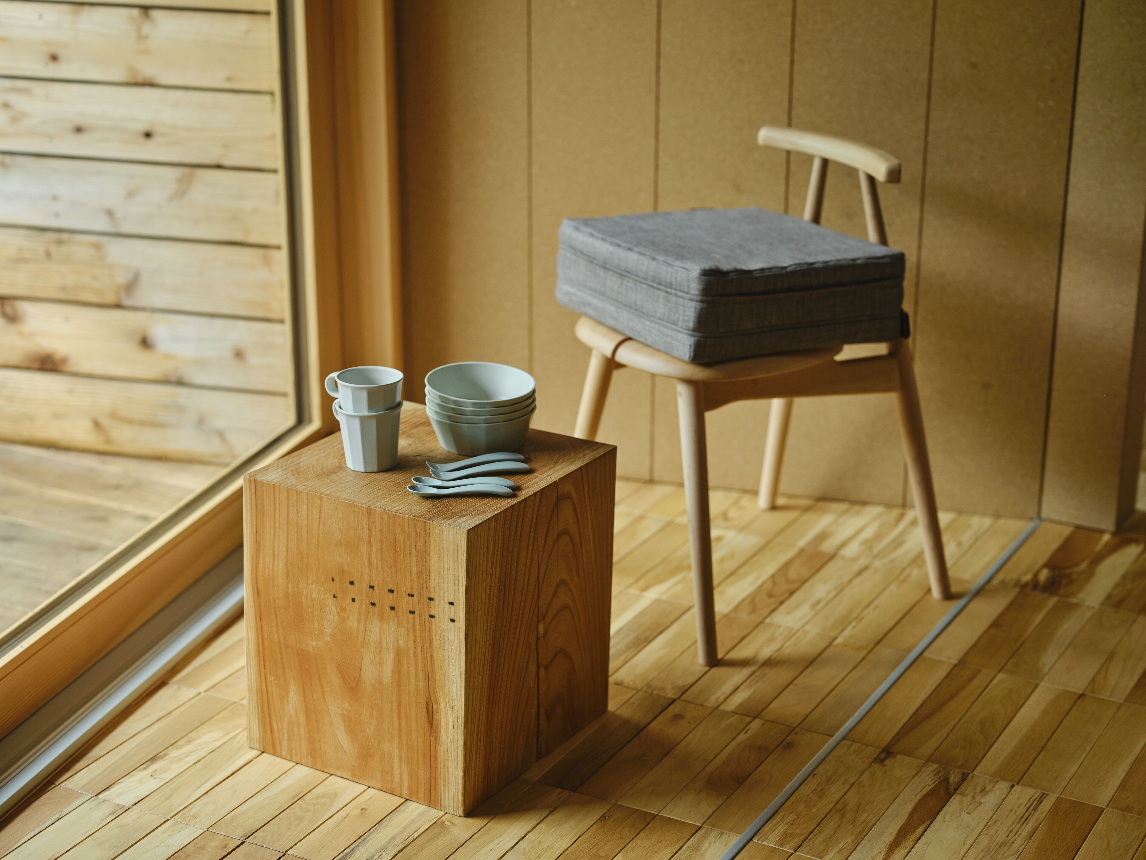 A wooden stool with a cushion, a stack of bowls, and a cup on a wooden platform.