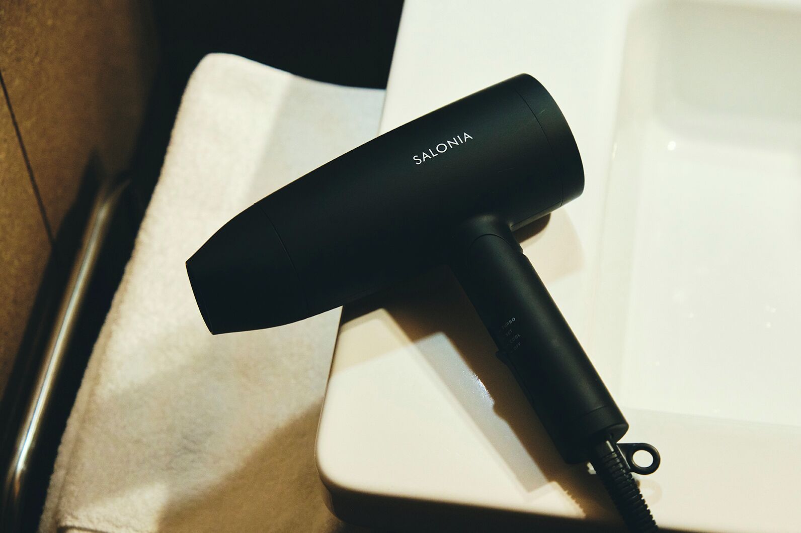 A black hairdryer with the brand name 'SALONIA' on it, placed on a white surface next to a beige towel.