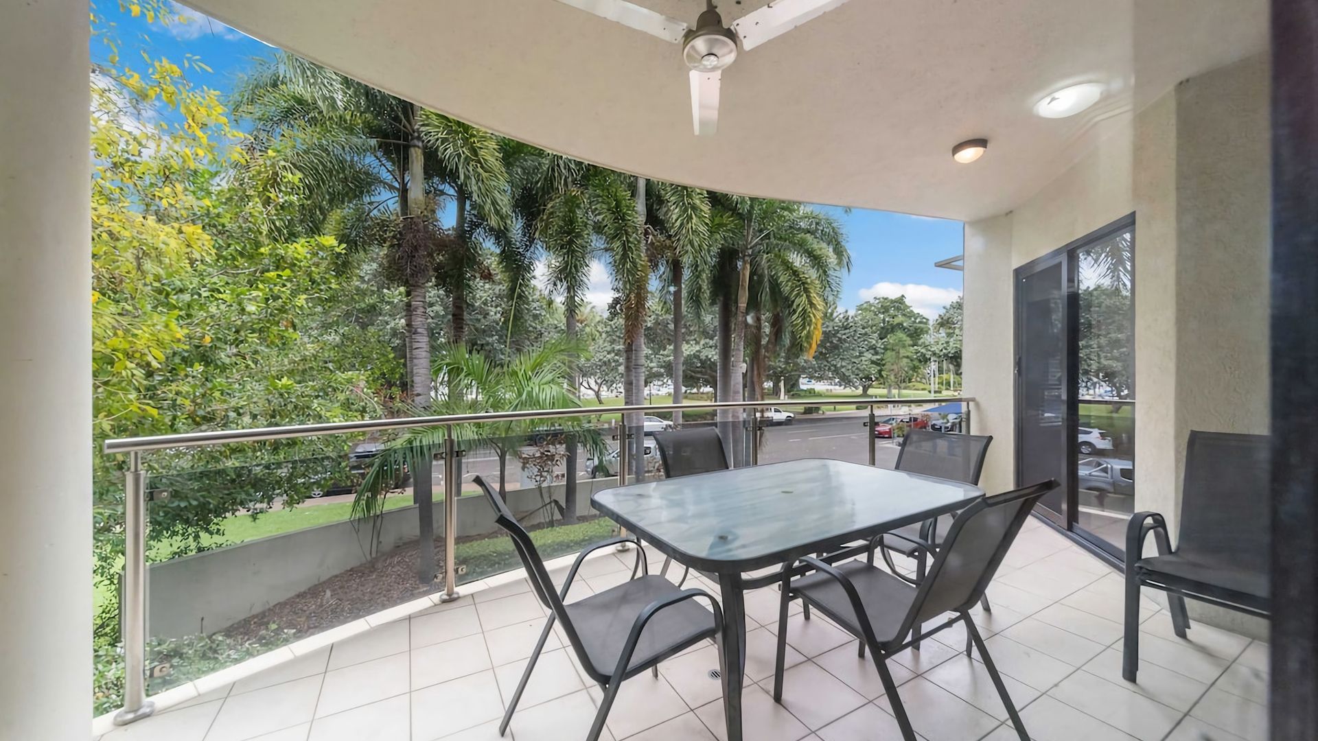 A balcony with a glass table and chairs, a ceiling fan, and a view of trees and a road.