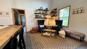 Un salon chaleureux avec une table à manger en bois, deux fauteuils bleus, un tapis à motifs et un banc. On peut également apercevoir des étagères ornées d’objets décoratifs et une fenêtre offrant une belle vue.