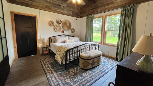 Une chambre à coucher avec un lit, un ventilateur de plafond, une table de chevet, une lampe, une fenêtre avec des rideaux et une vue sur l’extérieur.