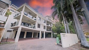 A multi-story residential building with balconies, a paved driveway, and a parking area.