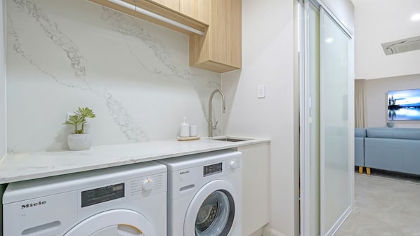 A compact laundry area with two Miele washing machines, a marble backsplash, a wooden cabinet, and a sliding glass door leading to a living area.