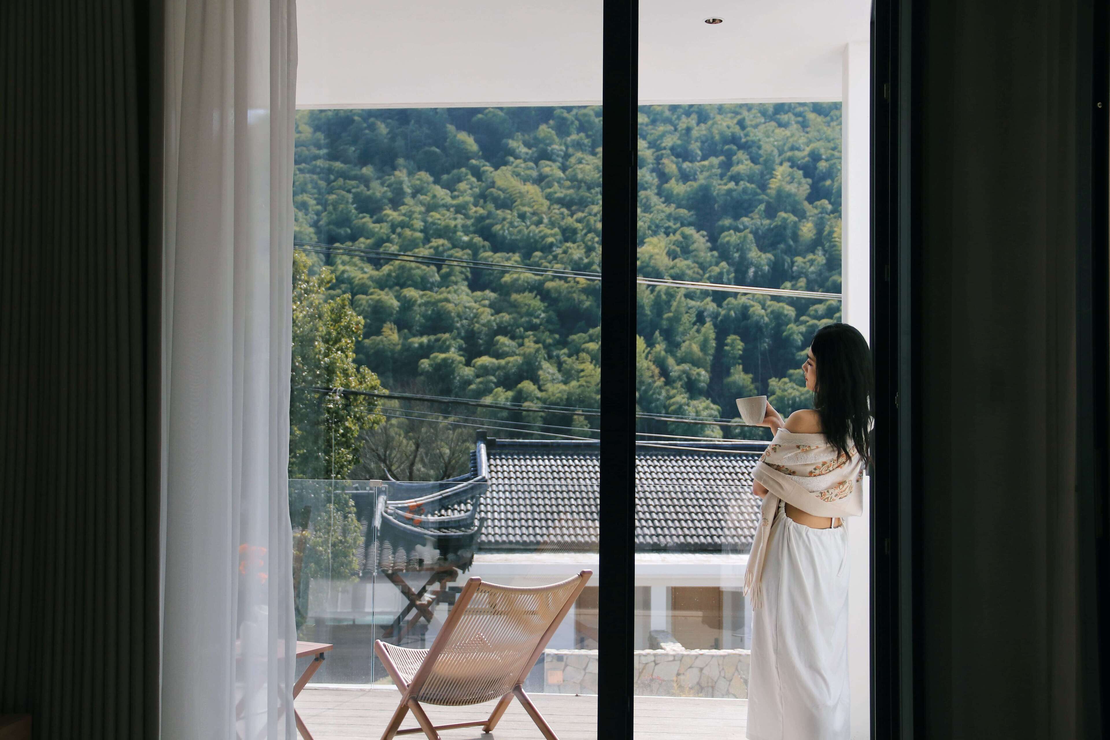 Ein Balkon mit Blick auf eine bewaldete Hügelslandschaft, ein Stuhl und eine Person mit einer Tasse.