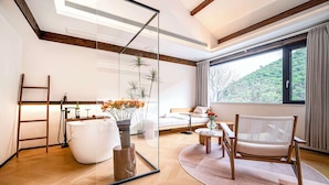 Un salon moderne avec une cloison en verre, une baignoire blanche, une chaise en bois, un canapé et une vue sur une forêt, visible à travers une grande fenêtre.