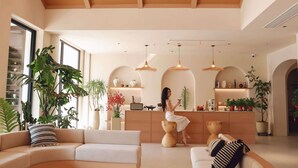 Un vaste espace de vie doté d’un haut plafond, de poutres en bois et d’un coin salon central avec un canapé et une table basse. On y trouve également plusieurs plantes en pot et, au fond, un coin cuisine.