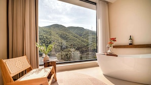 Une salle de bain moderne avec une grande baignoire, une chaise en bois, une petite table ornée d’un vase de fleurs et une vue sur une colline boisée.