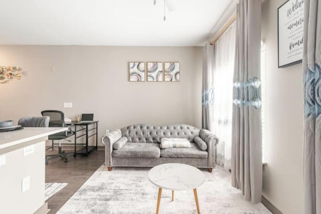 A living room with a grey sofa, a round coffee table, and a desk with a chair. There is a framed artwork on the wall and a window with curtains.
