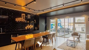 Un interior moderno con una larga mesa de comedor de madera, una pared de ladrillo negro con letreros de neón y grandes ventanales que ofrecen vistas al exterior.