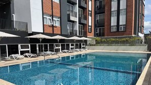 Una piscina con sillas de descanso y sombrillas, rodeada por un edificio moderno con balcones.