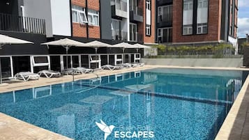 Een zwembad met ligbedden en parasols, omringd door een modern gebouw met balkons.