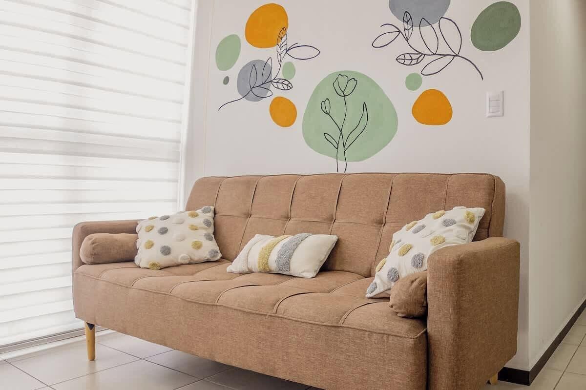 Ein beiges Sofa mit zwei gemusterten Kissen, eine Wand mit abstrakter Kunst und ein Fenster mit Jalousien.