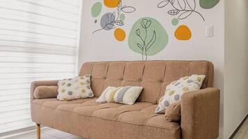 Ein beiges Sofa mit zwei gemusterten Kissen, eine Wand mit abstrakter Kunst und ein Fenster mit Jalousien.
