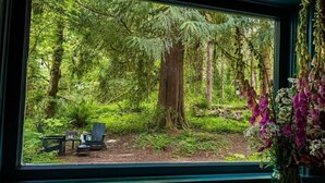 A window with a view of a forested area, a chair, and a vase with flowers.