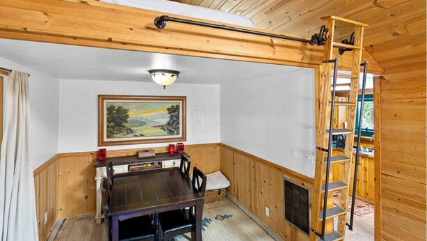 A wooden-paneled room with a dining table, chairs, a painting, a ceiling light, and a wooden ladder.