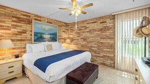 A bedroom with a brick wall, a bed with a blue blanket, a ceiling fan, and a nightstand with a lamp. - Oceanfront 3-Bedroom Condo w/ Direct Beach Access (South Padre Island)