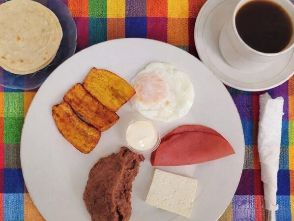 A plate with fried plantains, a fried egg, a slice of ham, a slice of cheese, and a small container of sauce, along with a cup of coffee on a colorful tablecloth. - Ecoturismo Monte Horeb (Gracias)