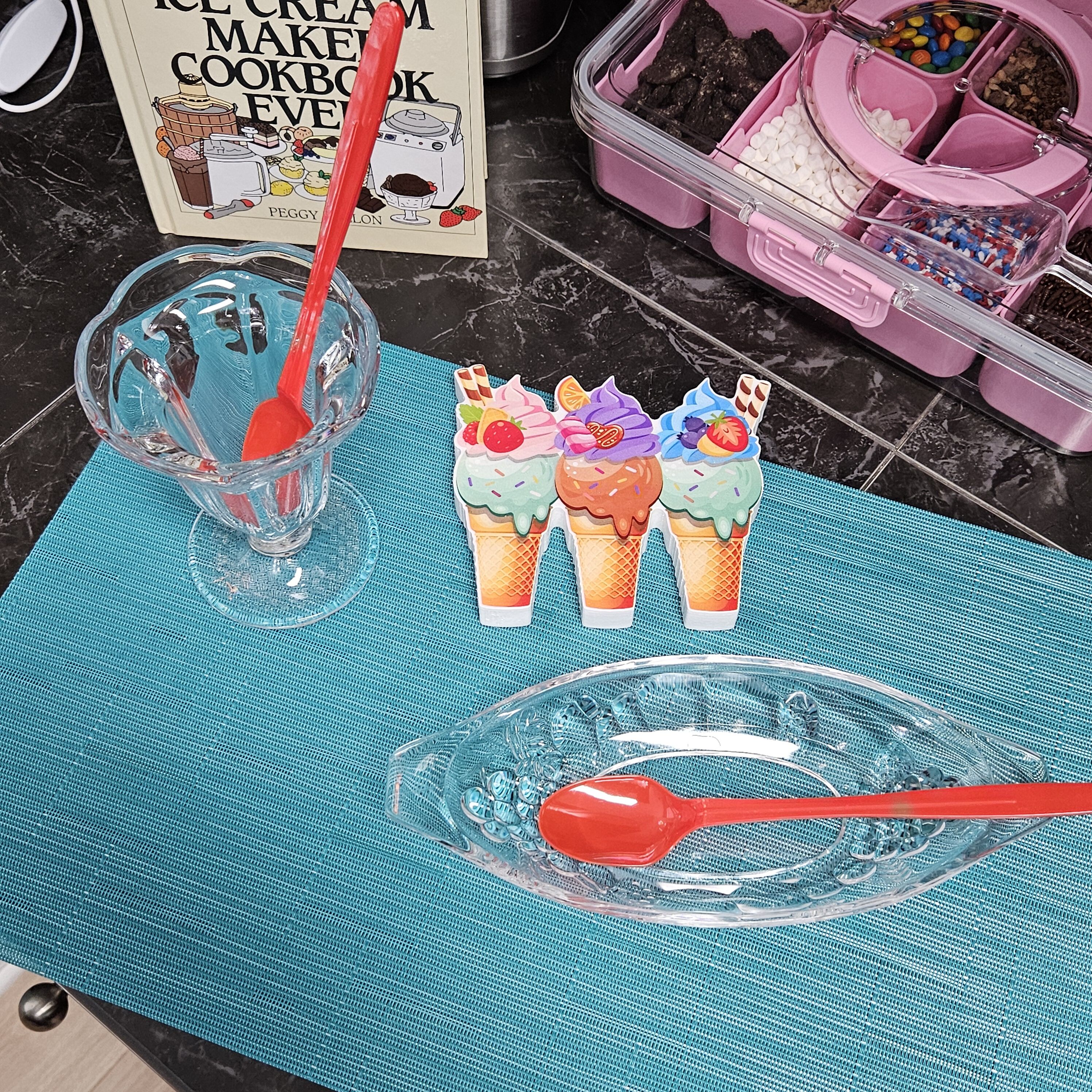 Un taulell de cuina amb un llibre de receptes, un recipient rosa ple de petits berenars i tres gelats.