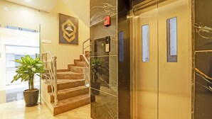 A hotel lobby with marble flooring, a staircase with metal railings, a potted plant, and a modern elevator with glass doors.
