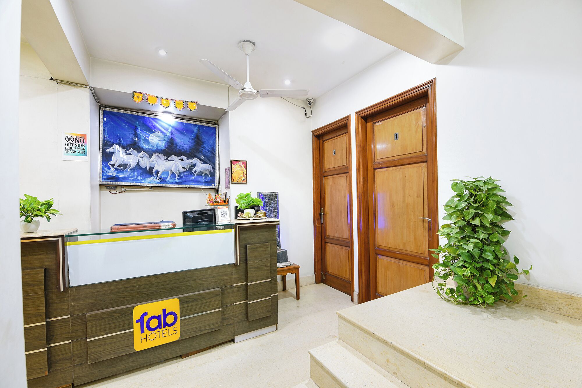 A hotel reception area with a wooden reception desk, a ceiling fan, a wall-mounted TV displaying a horse race, and a sign indicating the hotel name.