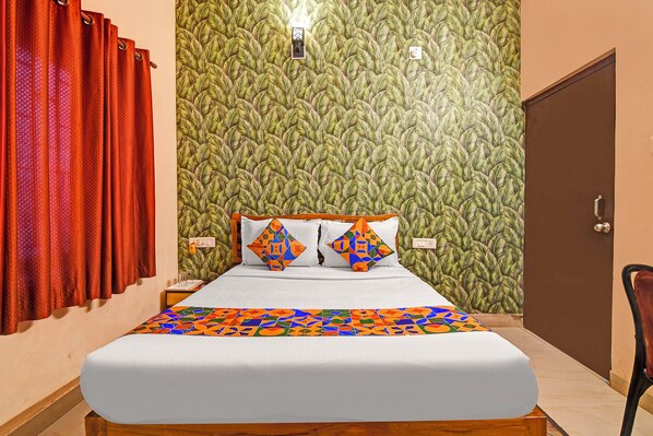 A hotel room with a bed, a wooden headboard, a wall with a leafy pattern, a door, and a red curtain.