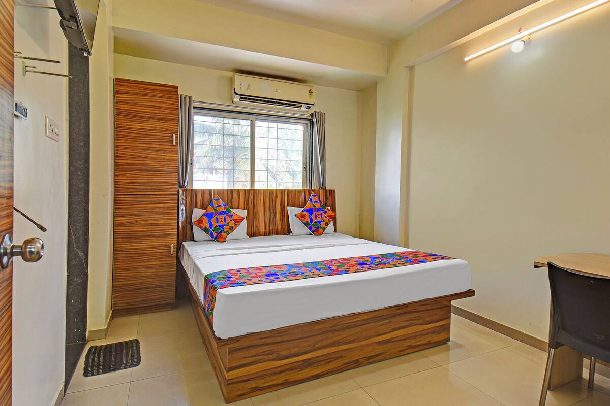 A bedroom with a wooden bed frame, a window with curtains, an air conditioning unit, and a small desk with a chair.