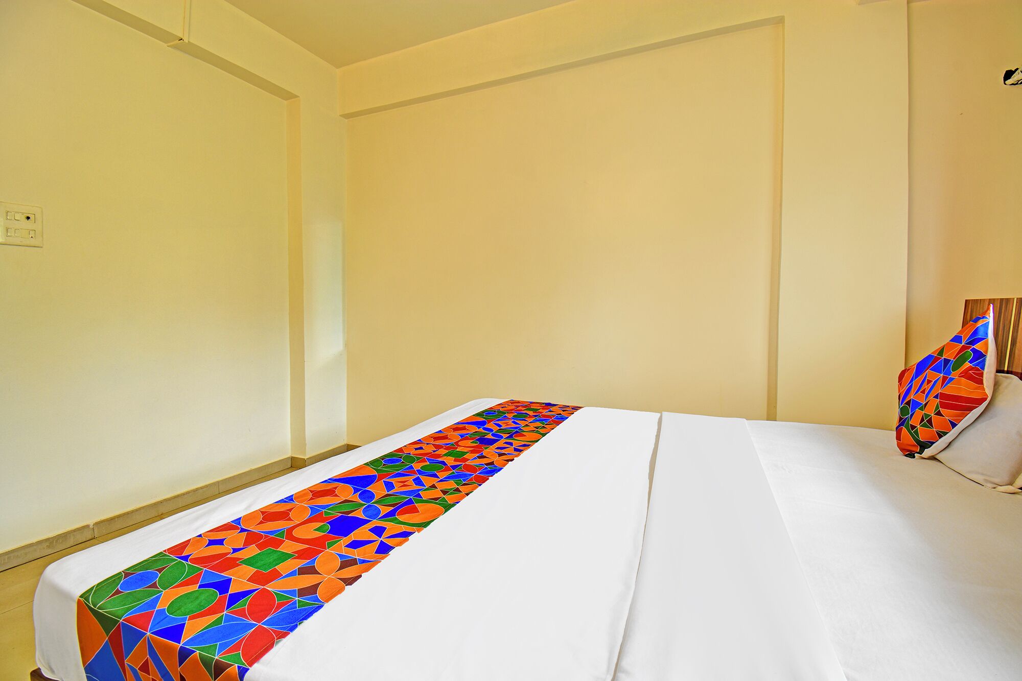 Une chambre avec un lit simple, recouverte d’une courtepointe blanche et agrémentée d’un tapis aux motifs géométriques colorés.