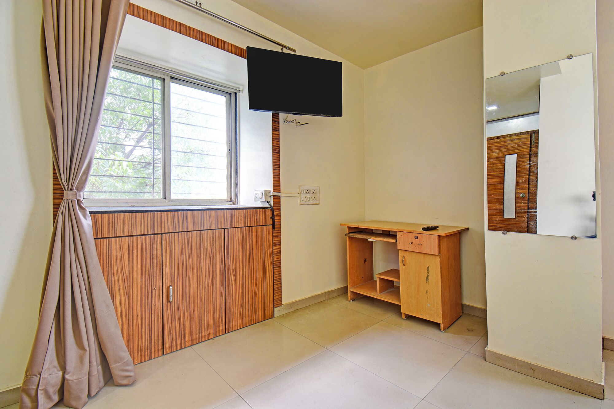 A room with a wooden wardrobe, a desk, a television mounted on the wall, and a window with curtains.