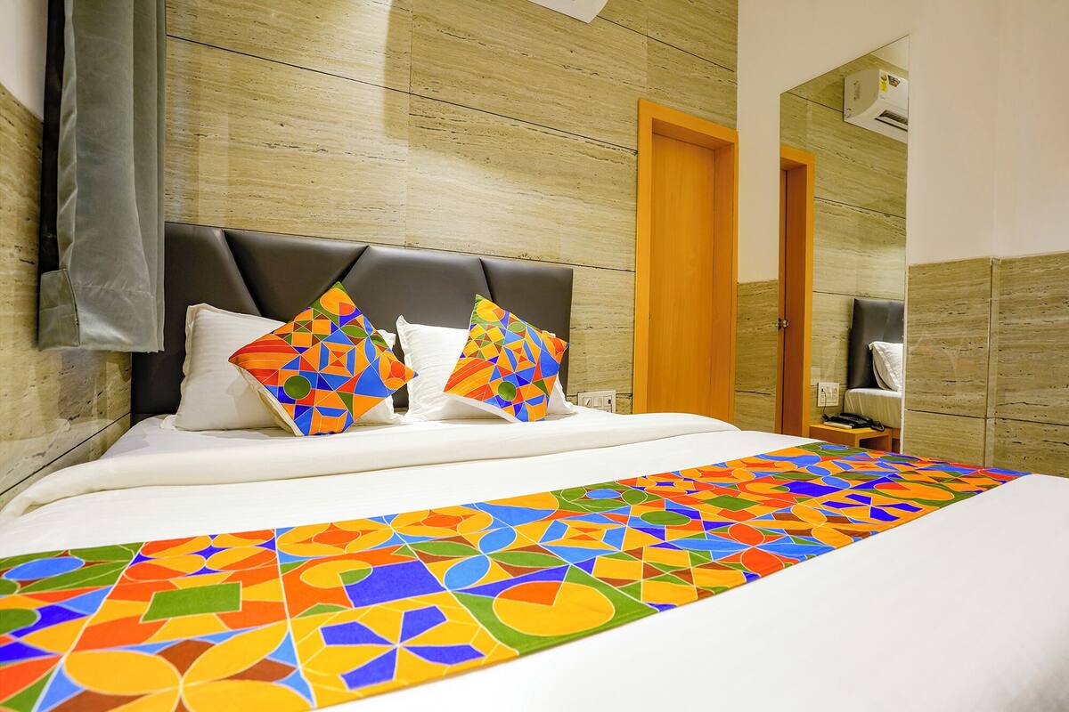 A hotel room with a bed, a wooden headboard, a colorful geometric throw blanket, and a mirror on the wall.
