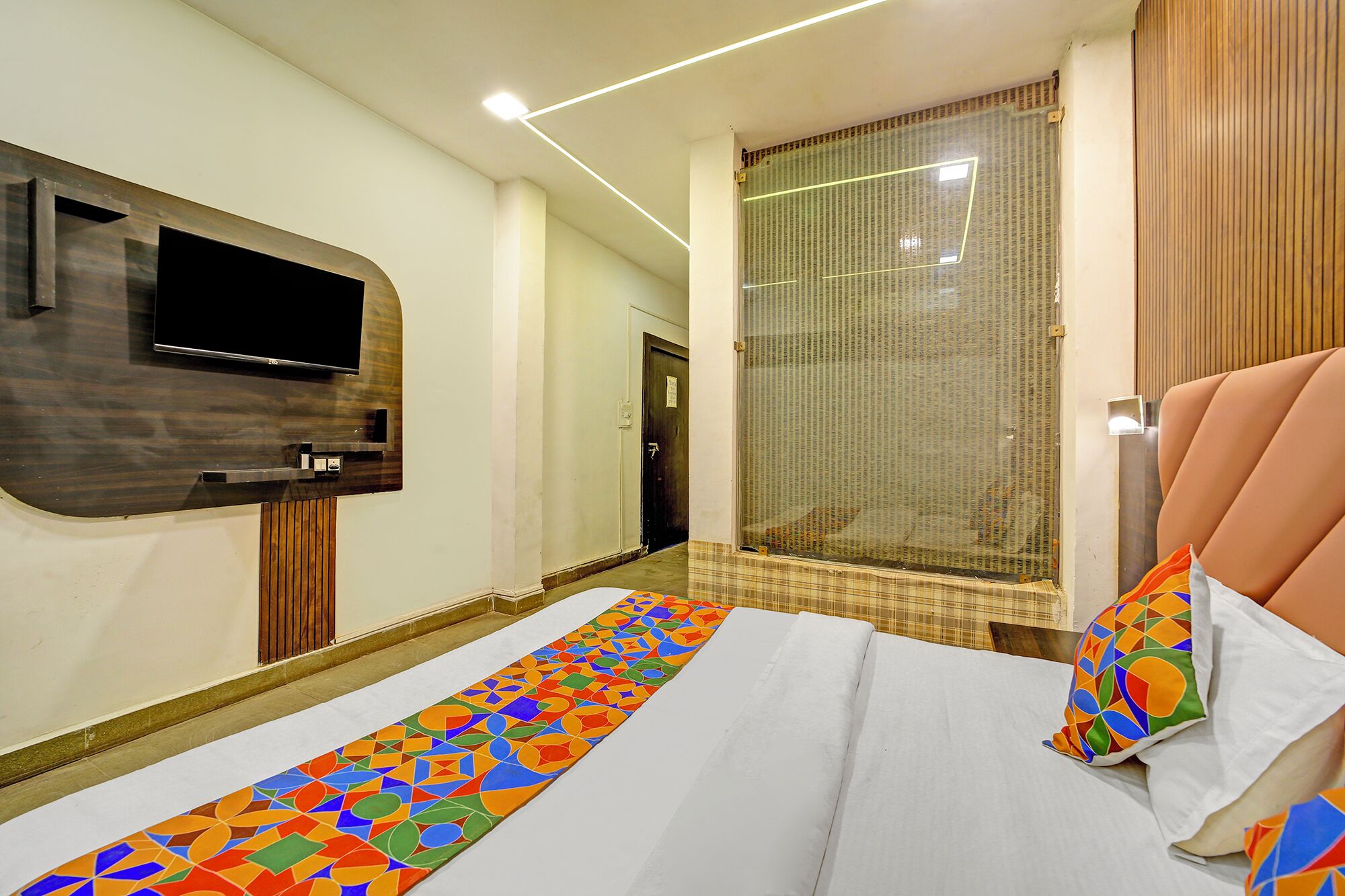 A hotel room with a large bed, a flat-screen TV mounted on a wooden panel, a shower area with a glass door, and a patterned rug.