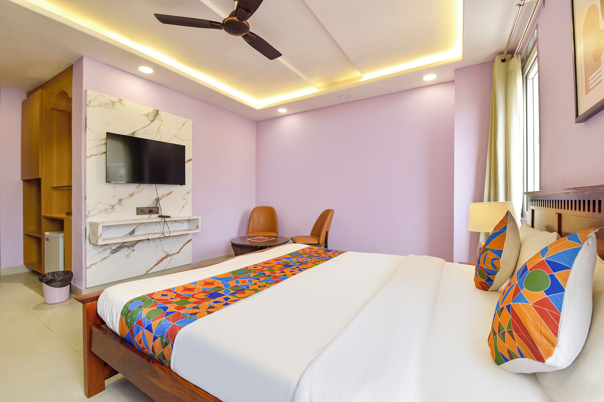 Habitación de hotel con cama, televisor montado en la pared, ventilador de techo y ventana con cortinas.