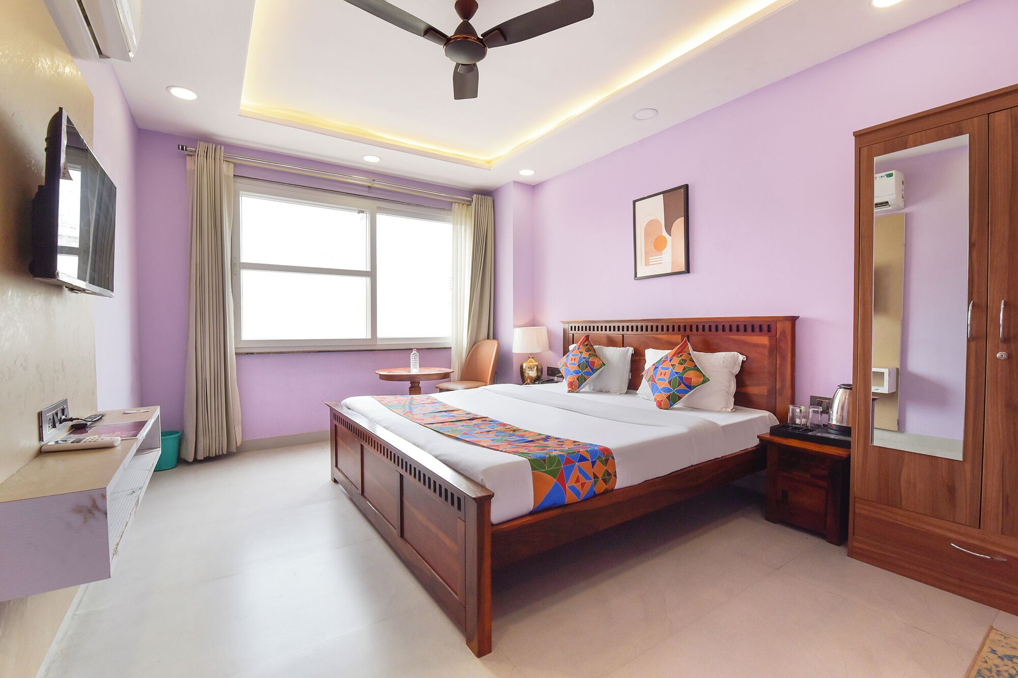 Habitación de hotel con cama de madera, un escritorio con silla, ventilador de techo, ventana grande con cortinas y armario.