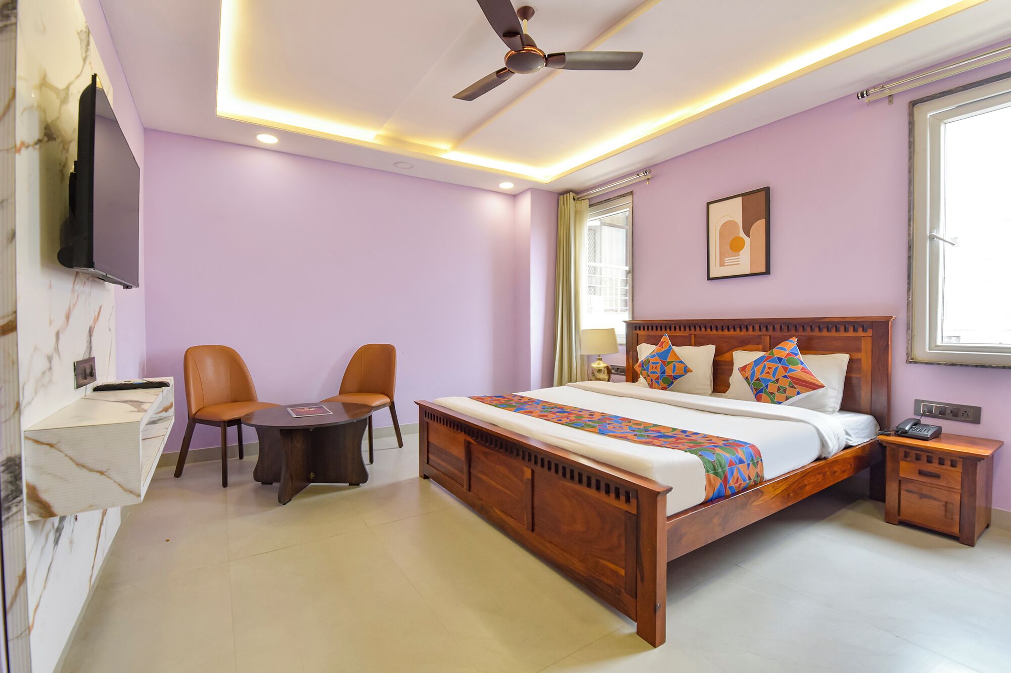 A bedroom with a bed, a desk, two chairs, a ceiling fan, and a TV.